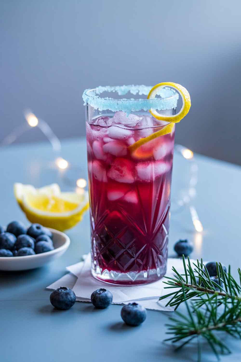 Vibrant purple-blue mocktail in a tall glass with crushed ice and a sugared rim, styled with blueberries and cool holiday lighting.