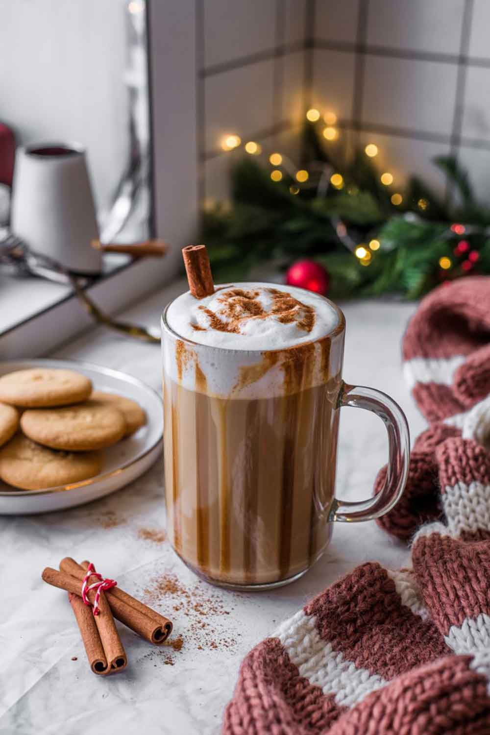 Spiked Chai Spiced Rum Latte A tall glass mug of spiked chai latte with frothed milk, cinnamon dusting, and a cinnamon stick garnish. Styled in a kitchen nook with a plate of spiced cookies, knit textures, and holiday garland.