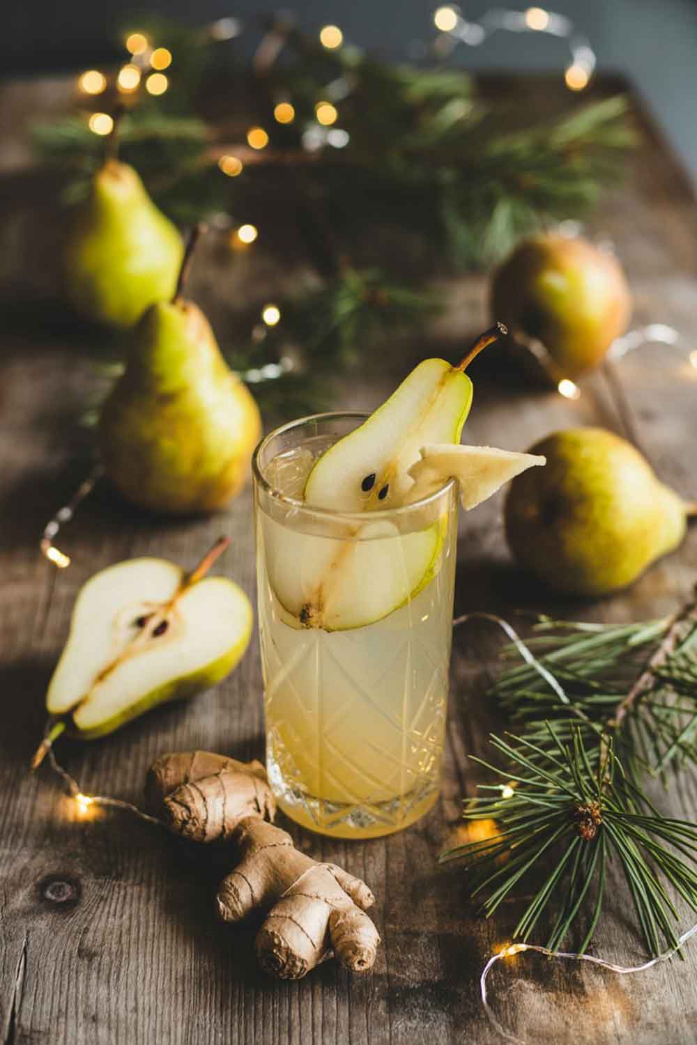 Spiced Pear Tequila Cooler A tall glass with a pale golden pear tequila cooler, garnished with a thin pear slice and ginger. Set among whole pears, pine branches, and warm golden string lights on a rustic winter table.