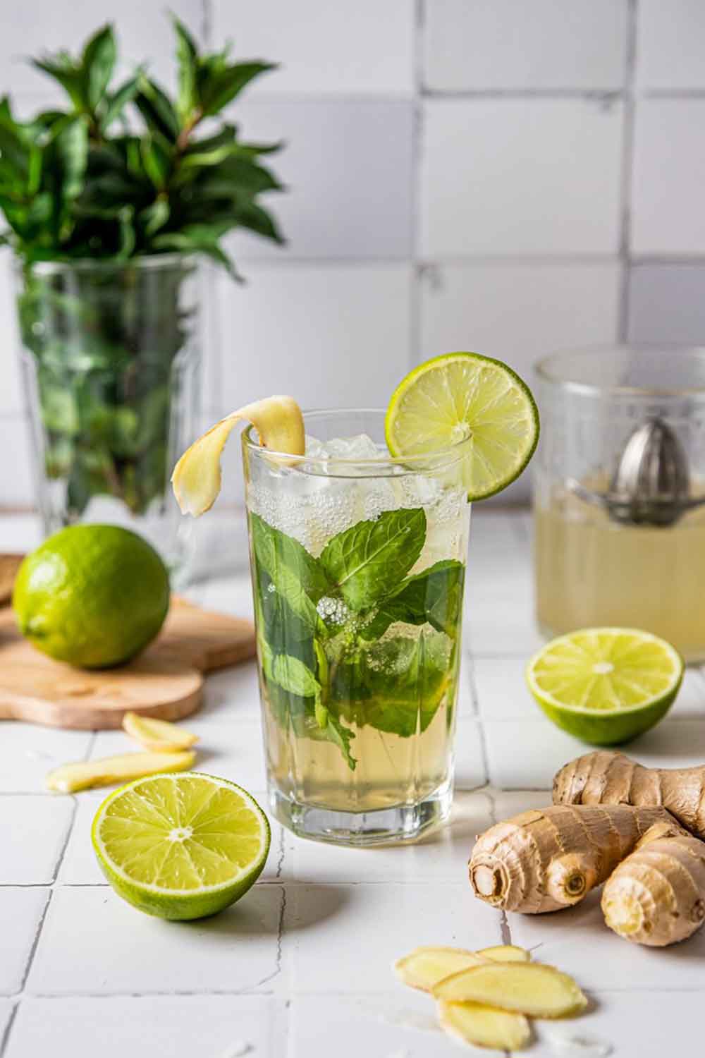 A sparkling mojito with muddled mint, ginger syrup, and lime juice in a clear highball glass. Fizzy, golden-green hue with crushed ice. Garnished with lime wheel and shaved ginger curl. Set on a white tile kitchen counter with fresh ginger root and a citrus juicer nearby.