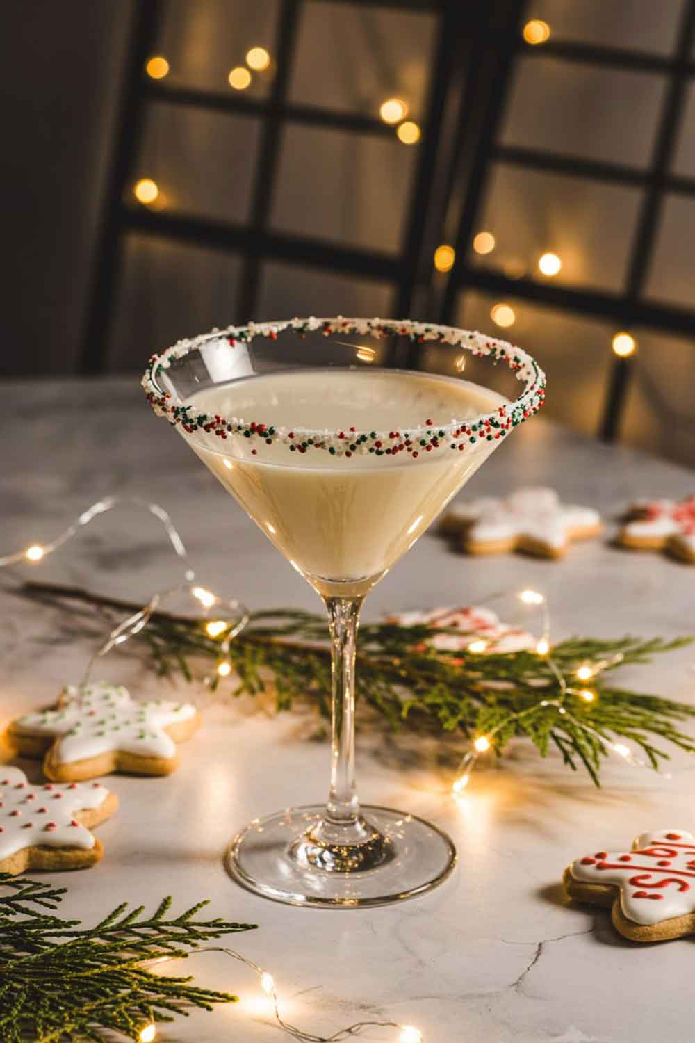 Sugar Cookie Tequila Martini A creamy vanilla martini in a martini glass with a sprinkle-covered rim. Styled on a festive table with sugar cookies, soft white lights, evergreen sprigs, and warm evening lighting. Cozy and playful Christmas theme.