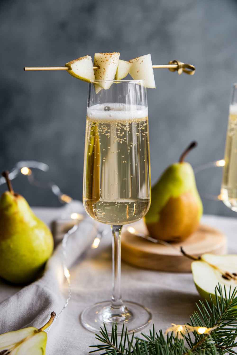 Pale golden pear spritz mocktail in a flute with a pear slice and nutmeg, styled with fresh pears and warm festive lighting.