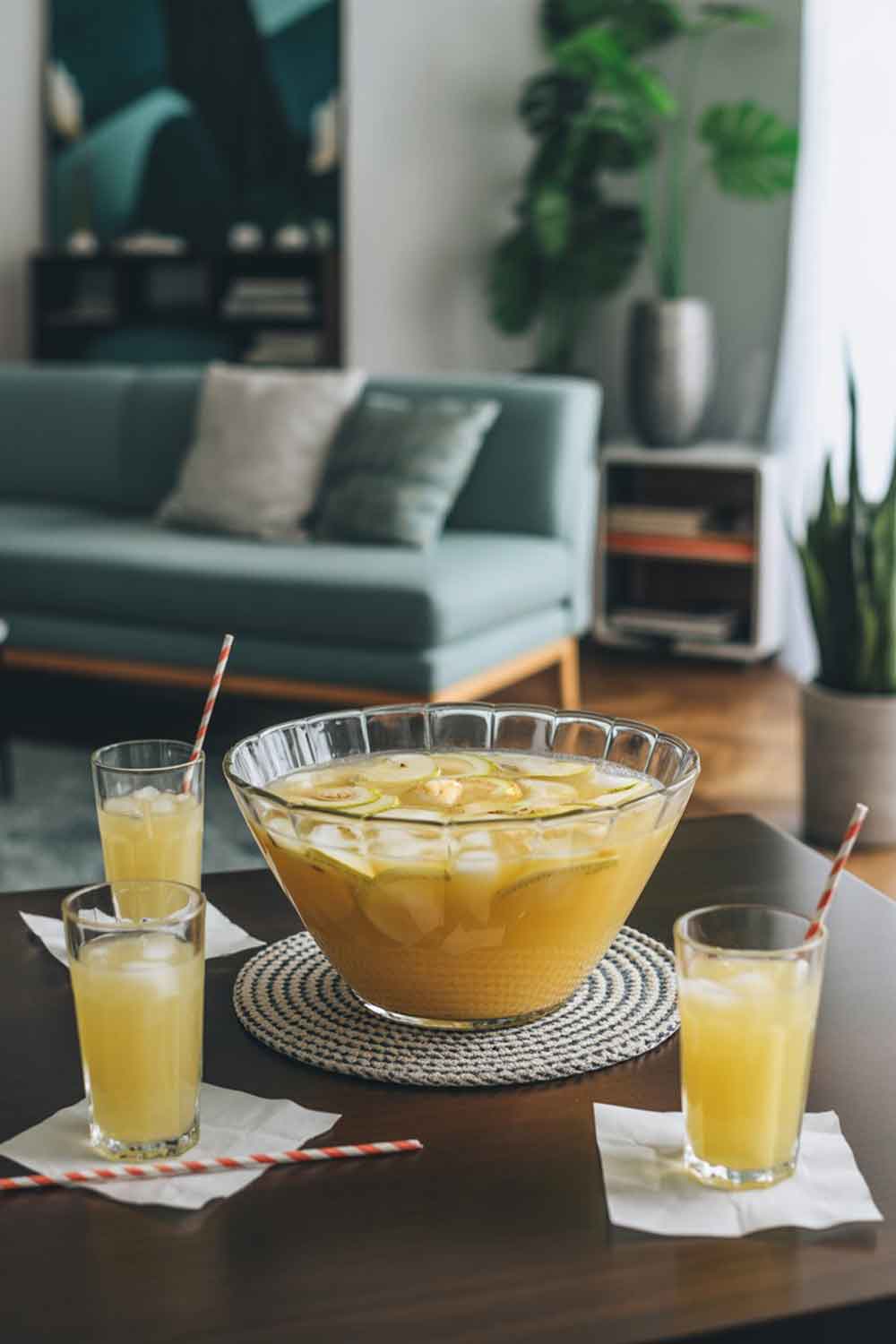 Pineapple Coconut Cream Punch Large punch bowl filled with pineapple coconut cream punch garnished with lime slices and ice, surrounded by tall glasses with striped straws on a coffee table.