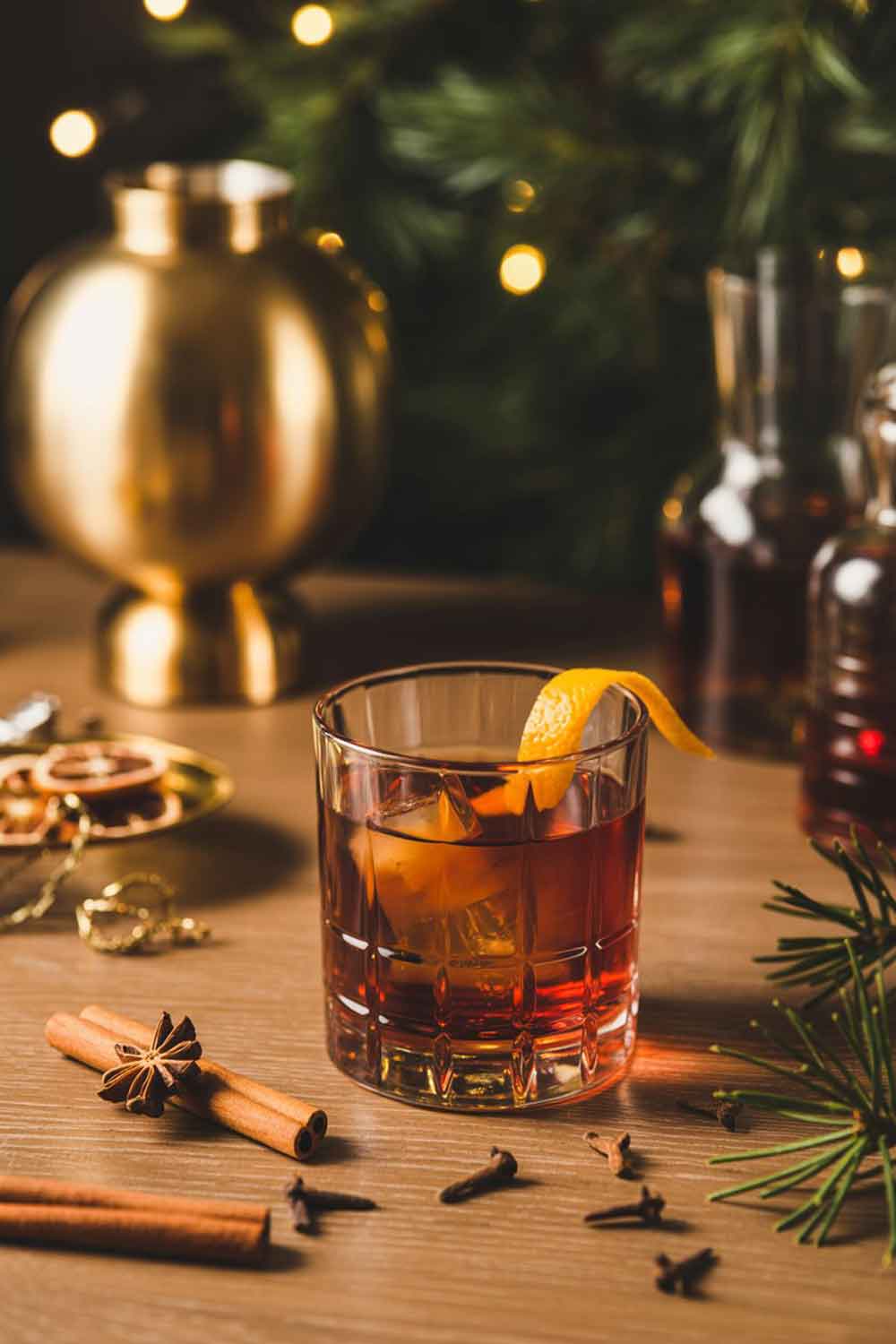 Gingerbread Tequila Old Fashioned A short heavy rocks glass holding a deep amber gingerbread tequila Old Fashioned, garnished with an orange peel twist. Christmas spices like cinnamon and cloves scattered on a wooden table, warm golden lamp light, pine branches, and subtle holiday décor in the background.