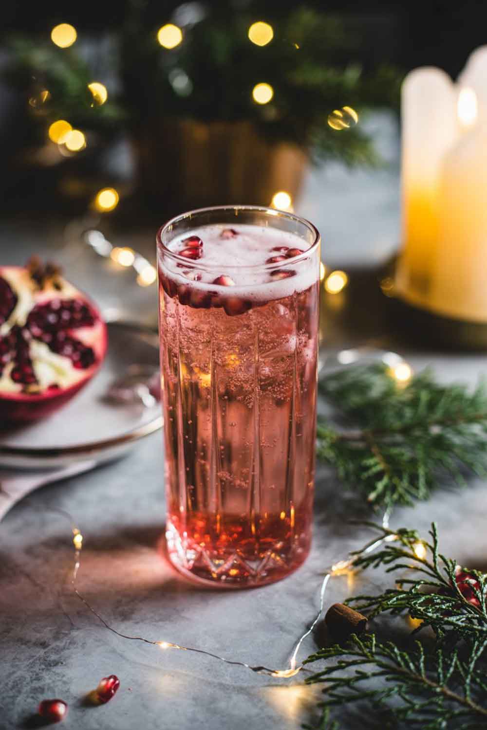 Pomegranate Tequila Fizz A tall glass with a fizzy pink-red pomegranate tequila cocktail, topped with bubbles and sprinkled with a few pomegranate seeds. Styled on a rustic table with soft fairy lights, evergreen sprigs, and a candle in the background for a warm evening glow.