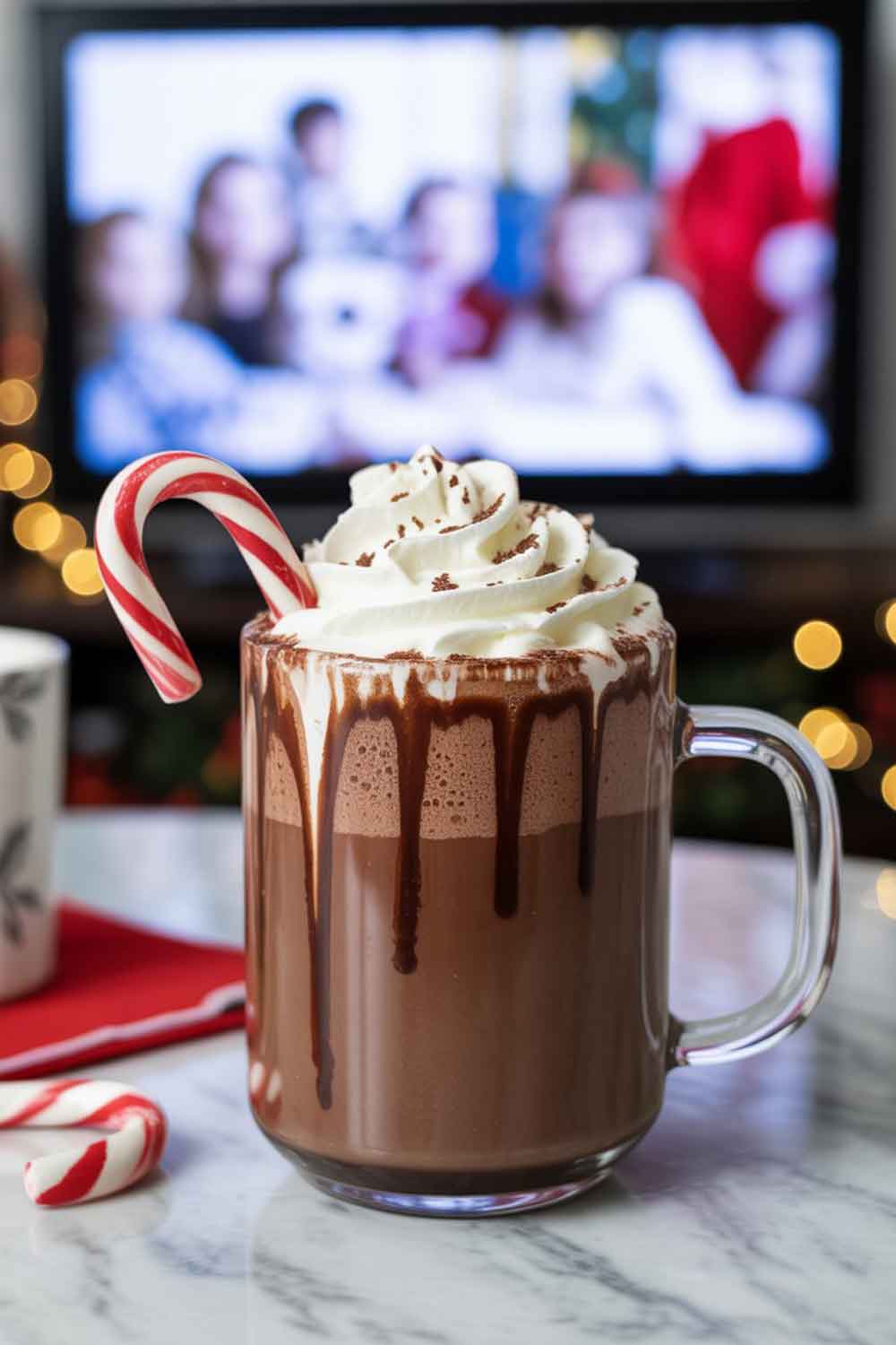 Gingerbread Rum Punch A festive mug of hot chocolate with whipped cream and a candy cane stir stick. Rich and creamy texture, slight chocolate drizzle down the side, served on a side table with a holiday movie on TV blurred in the background.