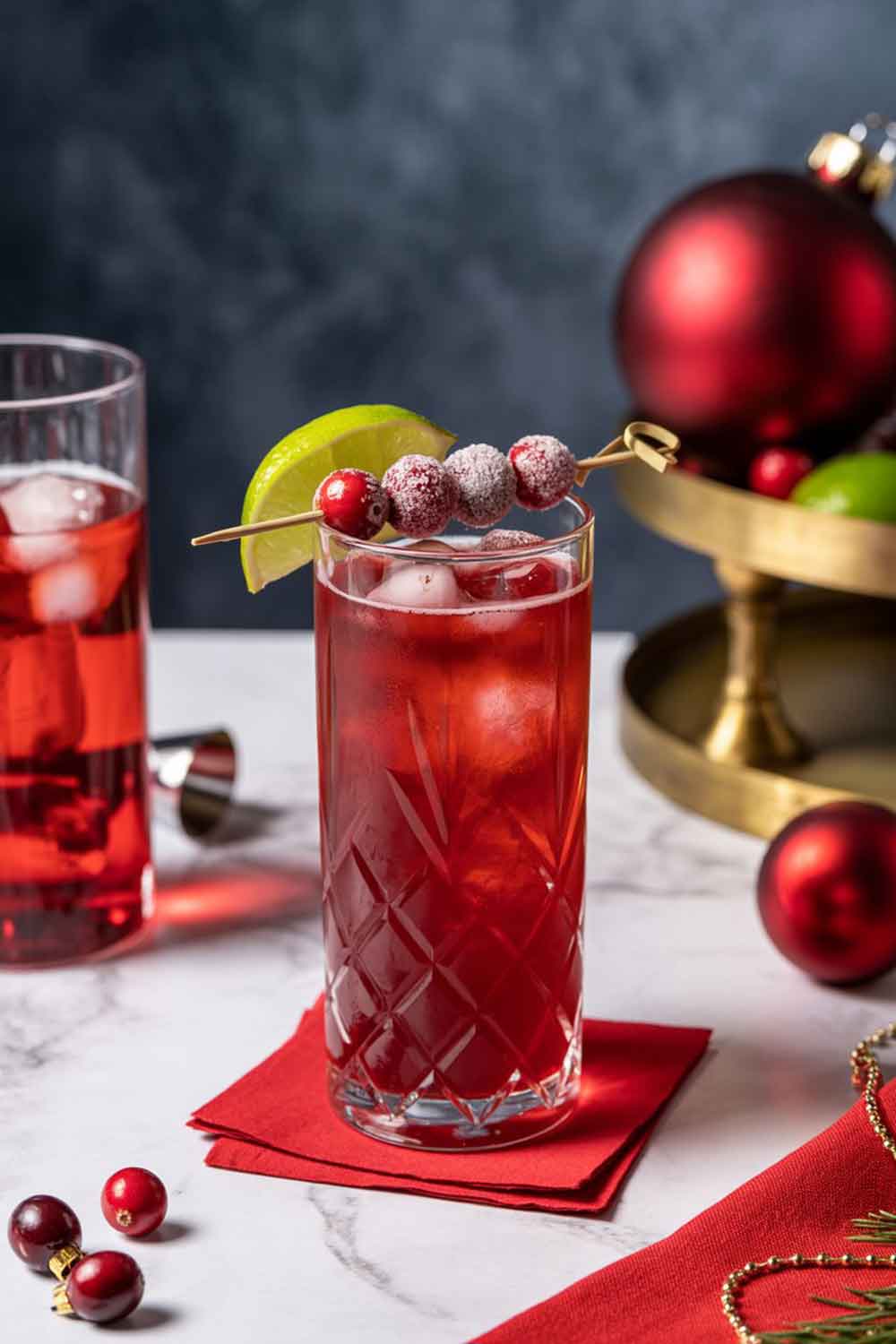 Cranberry Spiced Rum Fizz A tall highball glass of cranberry spiced rum fizz, deep red in color, garnished with sugared cranberries and a lime wedge. Styled with red ornaments, cranberries, and a festive napkin on a bar cart or dining table.