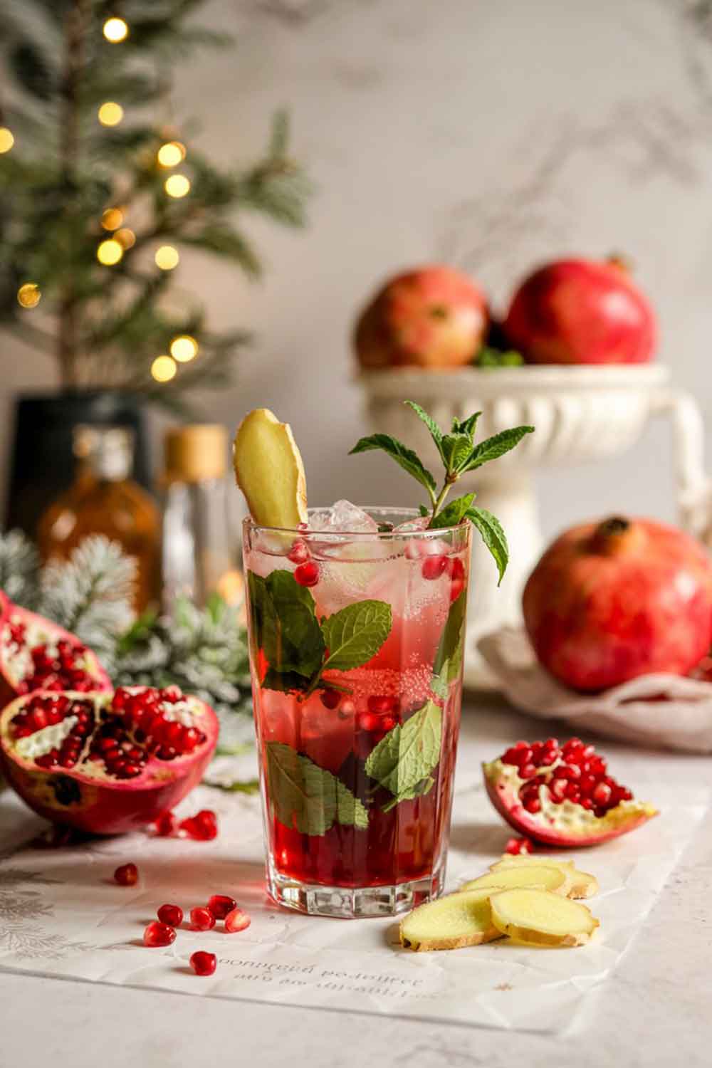 A tall mojito glass filled with deep pink pomegranate ginger mojito. Packed with crushed ice, muddled mint, and pomegranate juice, with ginger syrup swirled through. Garnished with pomegranate seeds and a thin slice of fresh ginger. Placed on a kitchen island with a bowl of pomegranates and winter greenery in the background.