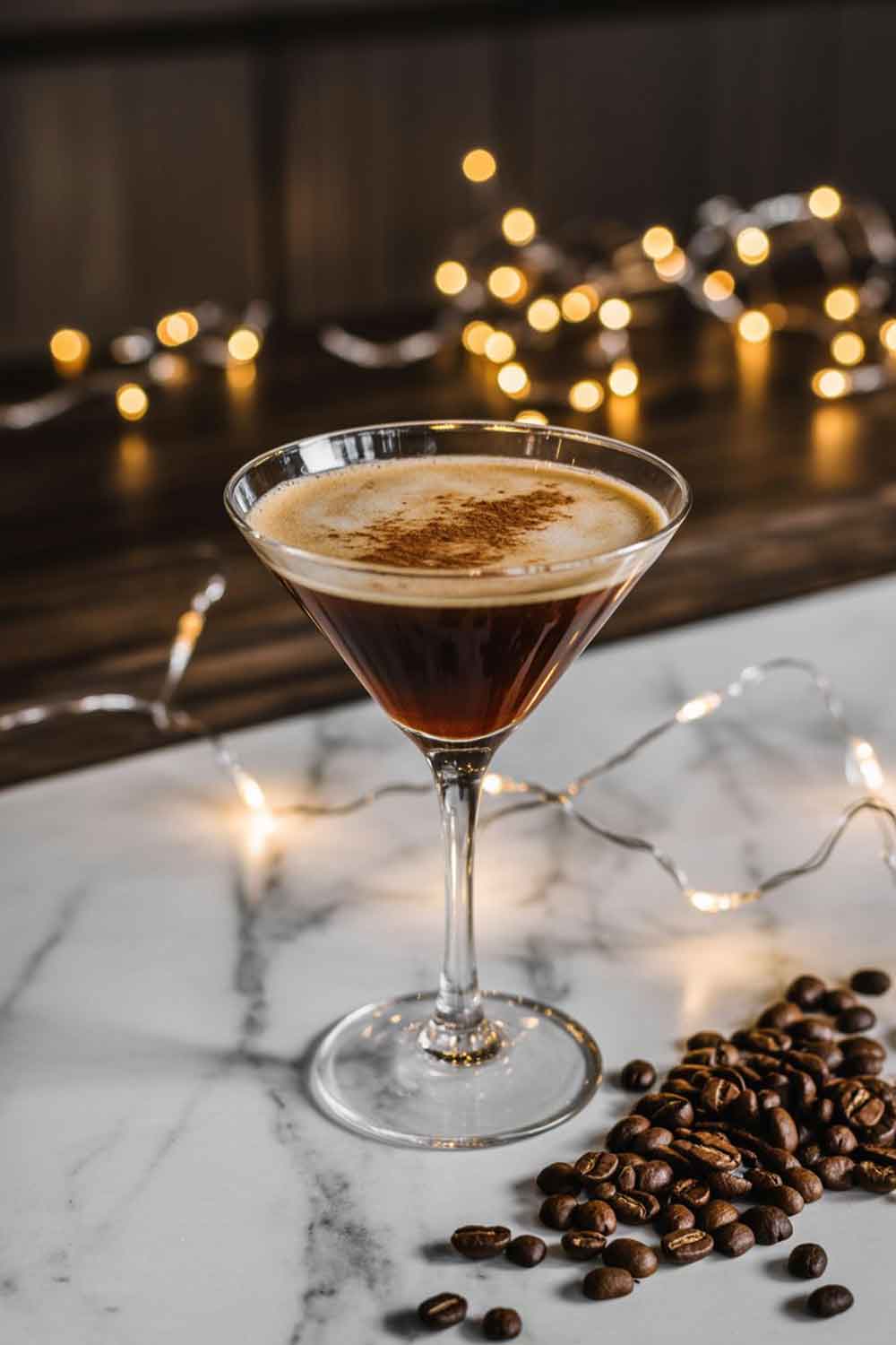 Spiced Rum Espresso Martini A sleek martini glass with a rich brown espresso martini topped with a sprinkle of cinnamon. Styled with coffee beans scattered nearby and soft golden Christmas lights twinkling in the background. Set on a marble or dark wood surface.