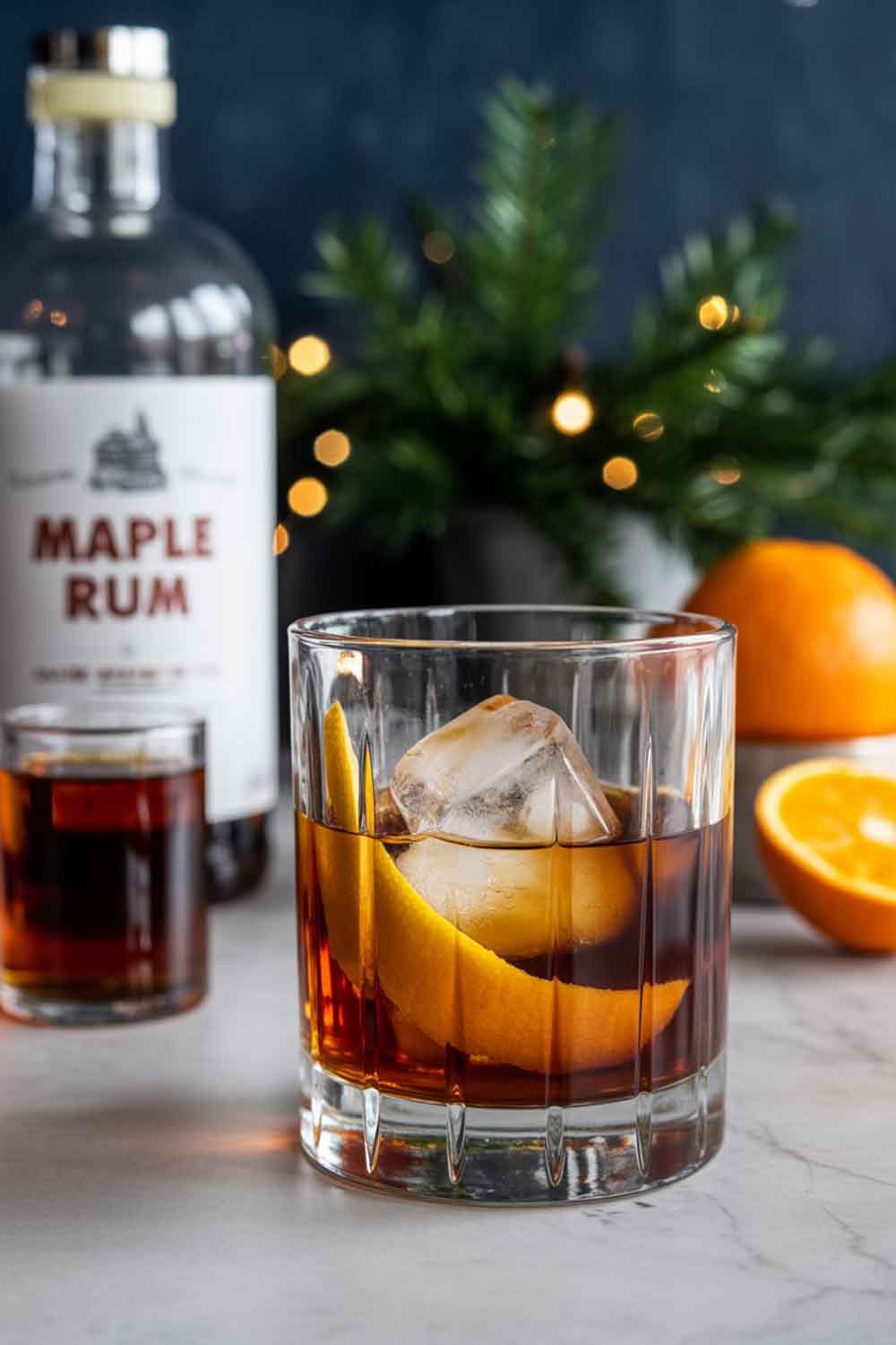 Maple Rum Old Fashioned A classic rocks glass with a maple rum old fashioned, garnished with orange peel and a large ice cube. Glass is slightly frosted. Styled with maple syrup bottle, holiday bitters, and oranges in a festive home bar setting.