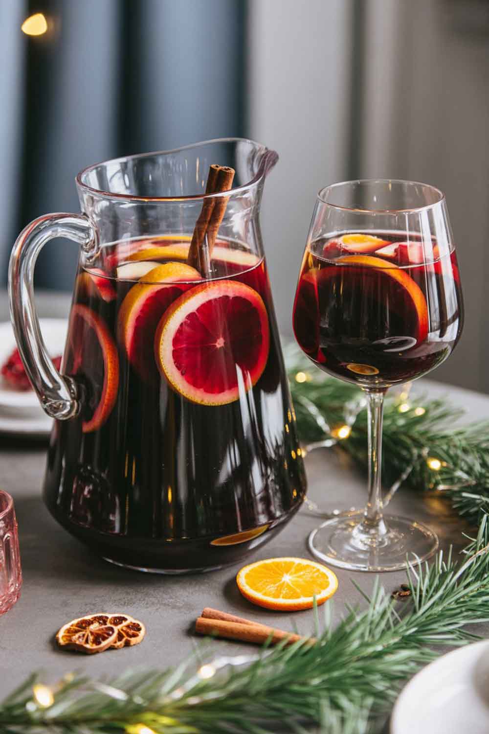 Tequila Christmas Sangria A large clear pitcher filled with deep red sangria, orange slices, apples, and a cinnamon stick, next to a matching filled wine glass. Arranged on a rustic holiday-themed table with greenery, string lights, and cozy winter décor.