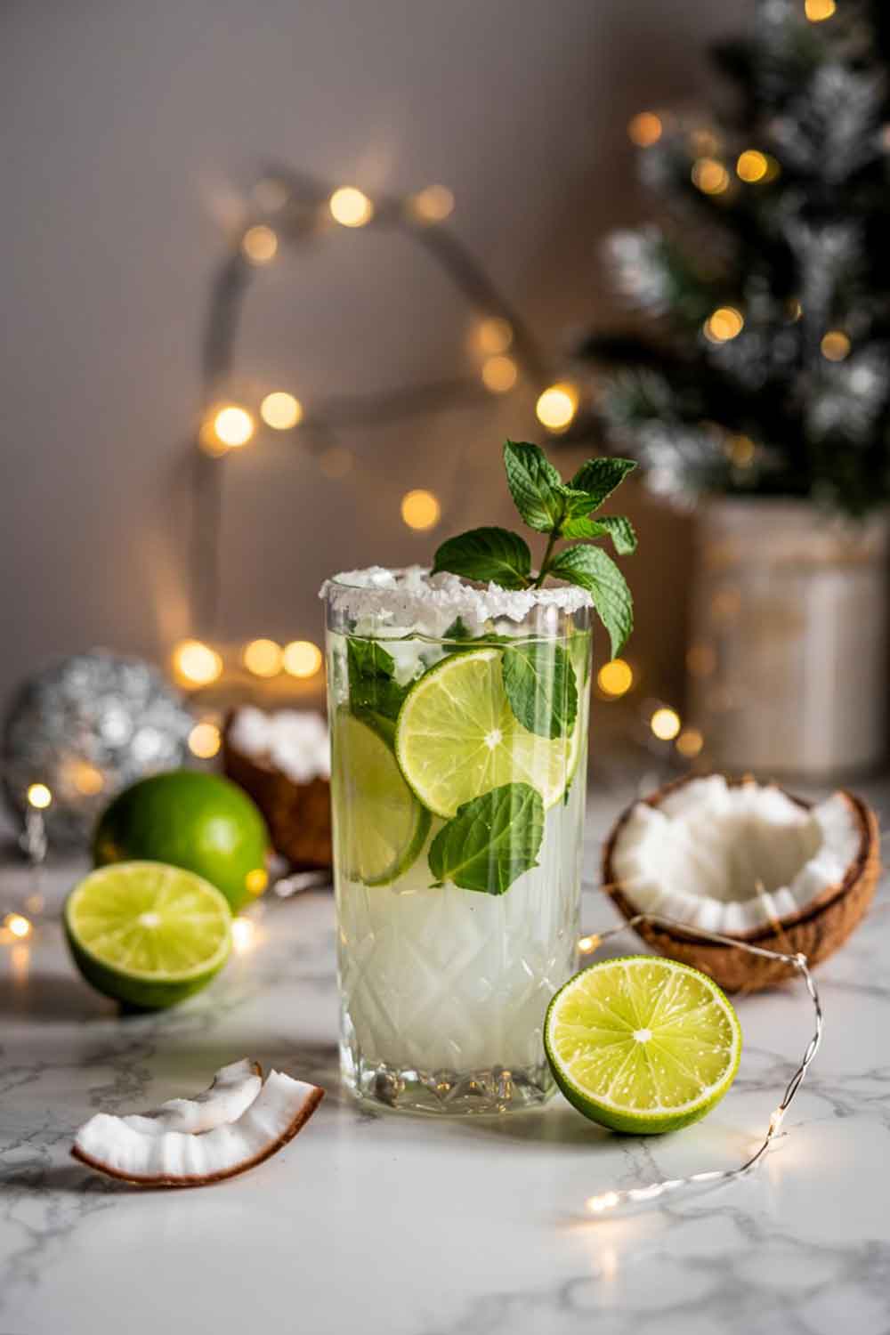 A creamy white coconut mojito in a clear highball glass with mint leaves and lime slices floating. Garnished with shredded coconut rim and a sprig of mint. Set on a marble kitchen surface with fairy lights and silver ornaments in the background.