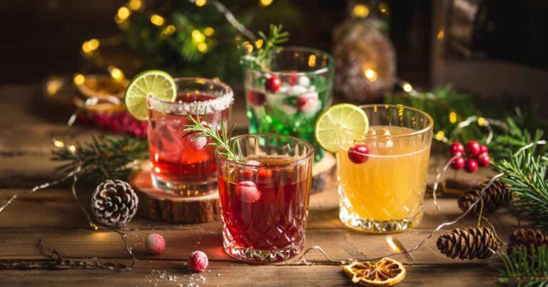 An assortment of Christmas vodka cocktails in festive glasses, featuring red, green, and golden drinks garnished with cranberries, lime slices, and rosemary, arranged on a rustic wooden table with pinecones, evergreen branches, dried citrus, and warm holiday lights in the background.