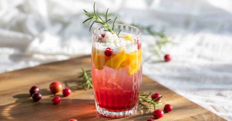 Layered pink and orange alcohol-free cocktail in a tall glass, topped with whipped foam, rosemary, and cranberries, styled on a wooden board in a warm home setting. A festive example of alcohol-free cocktails for Dry January.